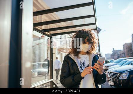 Junge schöne Frau trägt medizinische Gesichtsmaske an der Bushaltestelle in der europäischen Stadt stehen und halten ihr Handy. Stockfoto