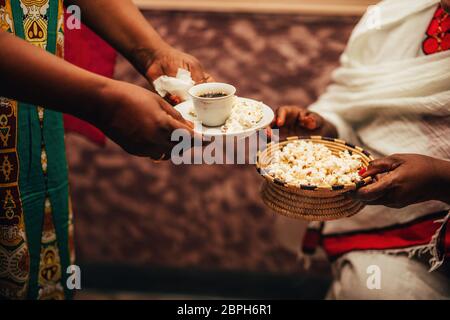 Afrikanische Frauenhände halten eine Tasse Kaffee und Strohschüssel mit Popcorn, eine traditionelle Art, Kaffee in Afrika zuzubereiten. Stockfoto