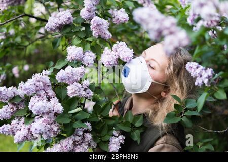 Junge blonde Frau riecht Flieder Baum Blüte durch FFP2 Atemschutzmaske. Konzeptionelles Bild der Quarantäne und Allergie des Corona-Virus. Stockfoto