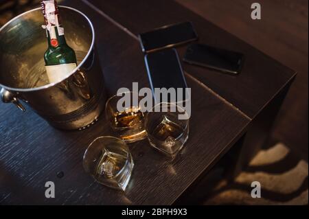 Auf einem dunkelbraunen Holztisch stehen drei Gläser Whiskey, ein Smartphone, ein Eiskübel und eine Flasche Whiskey. Stockfoto