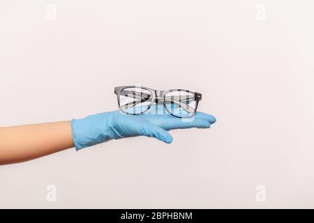 Profil Seitenansicht Nahaufnahme der menschlichen Hand in blauen chirurgischen Handschuhen halten und geben schwarze Brillen Rahmen. Indoor, Studio-Schuss, isoliert auf grauen Rücken Stockfoto