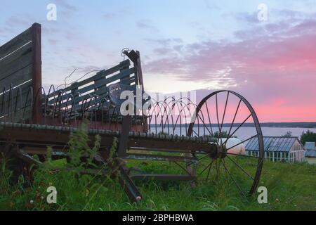 Vintage rostigen Heurachen mit einem alten Holzwagen auf dem Hintergrund eines dramatischen rosa bewölkten Himmel über dem Fluss bei Sonnenaufgang. Alte Landwirtschaft ausstattet Stockfoto