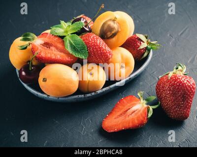 Detailansicht der Früchte und Beeren auf dunklem Hintergrund. Haufen von frischen Erdbeeren, Heidelbeeren, Aprikosen und Minze Blätter in trendigen Platte. Fokus auf stra Stockfoto