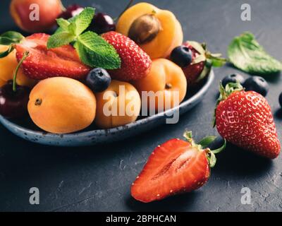 Detailansicht der Früchte und Beeren auf dunklem Hintergrund. Haufen von frischen Erdbeeren, Heidelbeeren, Aprikosen und Minze Blätter in trendigen Platte. Fokus auf stra Stockfoto