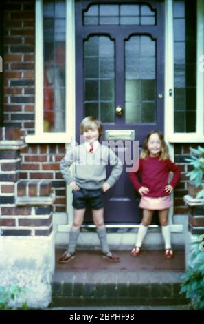 Ein junger Bruder und eine Schwester, lächelnd, stehen auf der Veranda ihres Familienhauses in ihren roten, grauen und weißen Schuluniformen in Vororten, Hatch End Park Estate, Milne Feid, Hatch End, Middlesex, Großbritannien Stockfoto