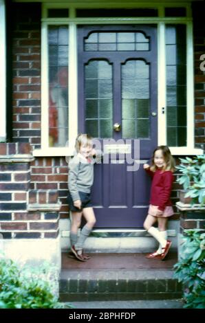 Ein junger Bruder und eine Schwester, lächelnd, stehen auf der Veranda ihres Familienhauses in ihren roten, grauen und weißen Schuluniformen in Vororten, Hatch End Park Estate, Milne Feid, Hatch End, Middlesex, Großbritannien Stockfoto