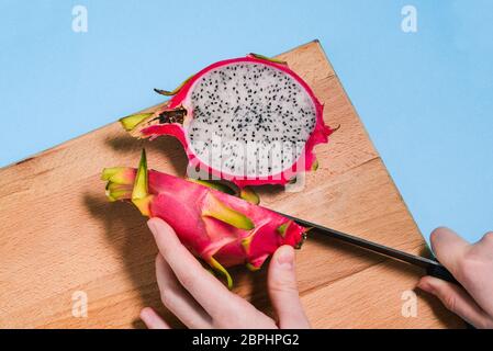 Schneiden Sie Pitaya Früchte. Nahaufnahme der weiblichen Hände, die Sommerdrachenfrucht schneiden. Reife Pitaya auf einem Holzbrett mit blauem Hintergrund. Stockfoto