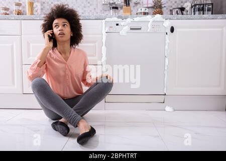 Traurige Frau ruft Techniker auf dem Handy zu fixieren Spülmaschine mit Schaum Aus Aus Stockfoto