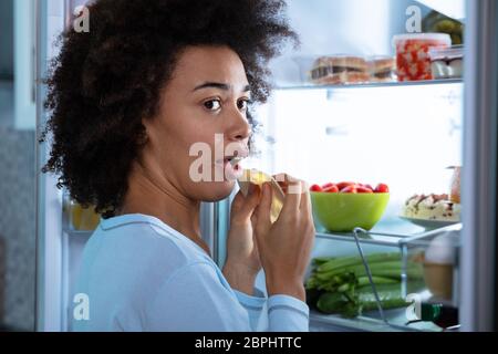 Junge afrikanische Frau Essen Scheibe Käse in der Nähe von offenen Kühlschrank Stockfoto