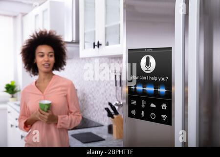 Junge Frau mit grünem Kaffee in der Hand auf Kühlschrank mit Spracherkennung Stockfoto