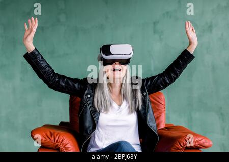 Senior und moderne Technologien. Schöne grau behaarte ältere Frau trägt schwarze Lederjacke, sitzt in bequemen roten Sessel in Stockfoto