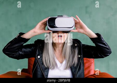 Virtual Reality Brille und ältere Frau. Aufgeregt überraschte ältere Dame mit geraden grauen Haaren, in schwarzen Lederjacke gekleidet, im roten Stuhl sitzend Stockfoto
