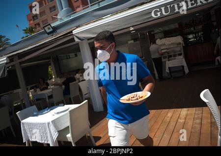 Während der teilweisen Sperrung im ganzen Land wird ein Kellner mit Gesichtsmaske gesehen, der während der teilweisen Sperrung auf der Terrasse der Strandbar "Gutierrez" einen Teller Garnelen an die Kunden serviert. Spanien durchläuft einen Plan zur Absenkung hin zu einer "neuen Normalität" durch Lockerung der Maßnahmen, die sich aus dem Ausbruch der COVID-19 ergaben. Während Phase 1 ist erlaubt, dass Menschen ihre Familien besuchen und mit einer Anzahl von maximal 10 Personen in Häusern, im Freien und Terrassen treffen können. Auch können Kirche, Geschäft, Geschäfte und Bars oder Restaurants mit ihren Terrassen auf 50% begrenzt wieder öffnen, die Sicherheit Abstand und die Verwendung von Face m halten Stockfoto