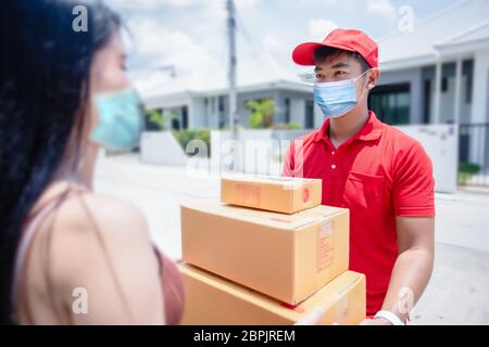 Asiatische Lieferung Soldaten tragen eine rote Uniform mit einer roten Kappe und Gesichtsmaske Handhabung Kartons, um die weibliche Kundin vor dem h zu geben Stockfoto