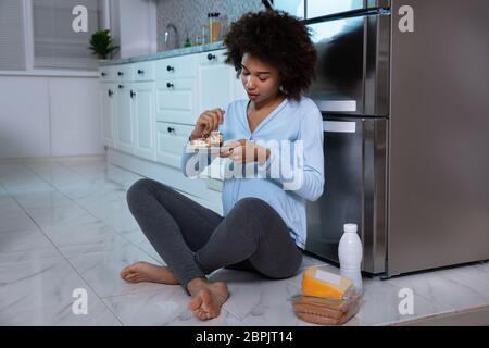 Junge schwangere Frau Essen Stück Kuchen sitzen vor dem offenen Kühlschrank in der Küche Stockfoto