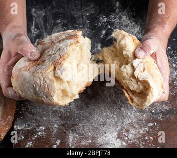 Männliche Hände Aufbrechen gebackenes Brot in die Hälfte über schwarzen Holztisch Stockfoto
