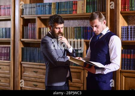 Intelligentes Elite- und wissenschaftliches People-Konzept. Männer im Anzug mit Vintage Bücherregalen auf Hintergrund. Professoren mit durchdachten Gesichtern lesen Buch in antikem Interieur. Intelligente Männer treffen sich in der Bibliothek. Stockfoto