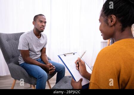 Junge afrikanische Mann sitzt auf Stuhl in der Nähe der Psychologin mit Zwischenablage Stockfoto