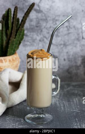 Dalgona Kaffee in einer transparenten Tasse mit einem Cocktail Stroh und Kaktus auf einem Betontisch. Stockfoto