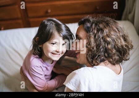 Junge Mutter und Tochter spielen im Schlafzimmer. Mama flüstert dem Kind ins Ohr, während sie beide lachen Stockfoto