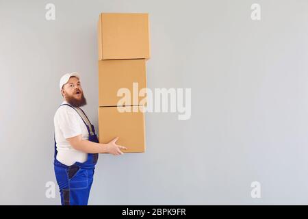 Lustige fette bärtige Lieferung Kurier Lader Mann in einer blauen Uniform mit einem Karton auf einem grauen Hintergrund. Stockfoto