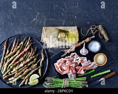 Speck eingewickelt Spargel im Ofen mit Knoblauch und Meersalz gebacken serviert auf einem schwarzen Teller mit Olivenöl auf einem dunklen Beton Hintergrund, Draufsicht, Fla Stockfoto