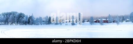Sehr breiter Panoramablick auf schwedisches Dorf mit roten und gelben Holzhäusern im Winter bedeckt auf gefrorenen Flussküste am Fichtenwaldrand, Lappland, Stockfoto