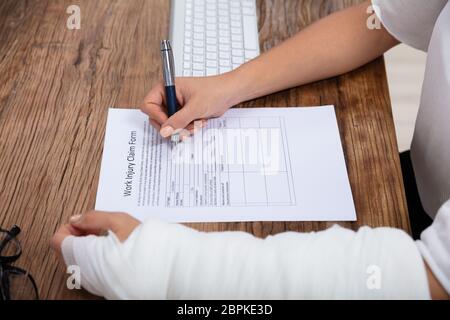 Nahaufnahme der Geschäftsfrau mit weißen Bandagen Hand Befüllen Arbeitsunfall Antragsformular auf hölzernen Schreibtisch Stockfoto