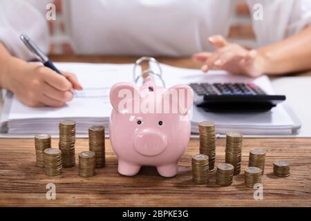Gestapelte Münzen und rosa Sparschwein Vor Geschäftsfrau Berechnung Rechnung auf hölzernen Schreibtisch Stockfoto