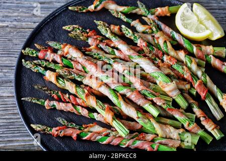 Ahornspeck umwickelt Spargel mit Knoblauch und Meersalz gegrillt serviert auf einem schwarzen Teller mit Zitrone auf einem alten Holzhintergrund, Draufsicht, Nahaufnahme Stockfoto