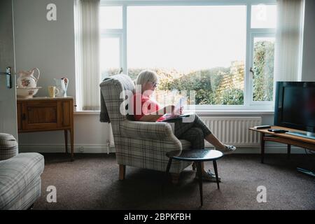 Eine ältere Frau sitzt in einem Sessel im Wohnzimmer ihres Hauses, Lesen durch Schreiben. Stockfoto