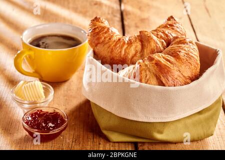 Frisch gebackene Croissants flockig in Stoffbeutel mit Butter, Marmelade und Kaffee auf rustikalen Holztisch serviert. Stockfoto