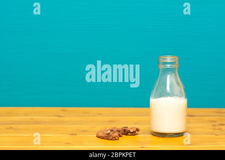 Ein Drittel pint Glas Milch Flasche halb voll mit frischen cremige Milch und ein halbes gegessen Chocolate Chip Cookie, auf einen hölzernen Tisch gegen eine blaugrüne Hintergrund Stockfoto