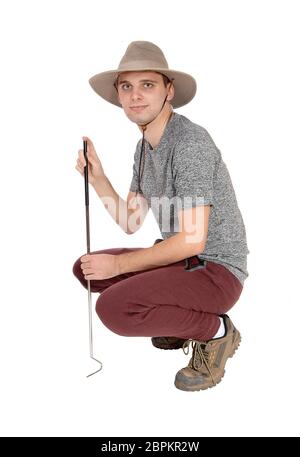 Ein schlanker junger Mann in einem grauen pullover mit Safari Hut und ein Werkzeug, um einige Baumstümpfe hocken auf dem Boden, auf weißem Hintergrund Stockfoto