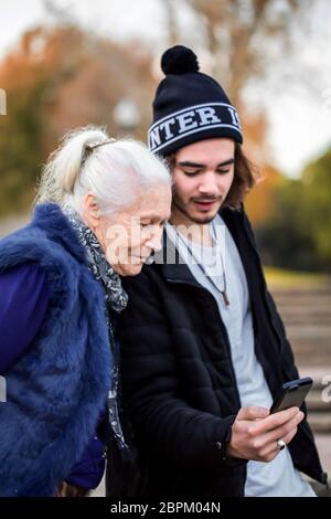 Der sympathische junge Mann erklärt seiner eleganten grauhaarigen Großmutter, wie man ein Smartphone benutzt. Sie laufen im Herbstpark und tragen lässige Kleidung. Stockfoto