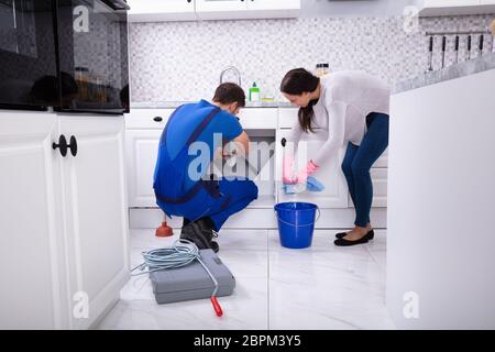 Junge Frau, Reinigung, während männliche Klempner Instandsetzung Waschbecken Rohr in der Küche Stockfoto