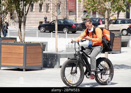 Belgrad, Serbien - 5. Mai 2020: Ein junger Kurier der Stadt fährt mit einem Elektrofahrrad auf dem Stadtplatz und schaut dabei auf das Mobiltelefon Stockfoto