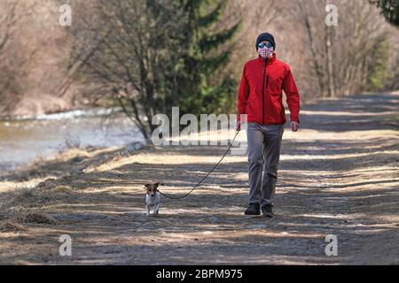 Junger Mann in roter Jacke, Mütze und Sonnenbrille, trägt hausgemachte Virus Gesichtsmaske, geht Hund auf Landstraße. Masken sind obligatorisch außerhalb des Hauses während Stockfoto