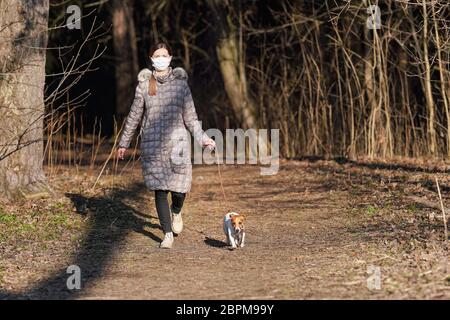 Junge Frau in warmer Jacke, trägt Virus Gesicht Mundmaske, geht ihren Hund auf Landstraße. Masken sind bei Coronavirus-Covid- Stockfoto