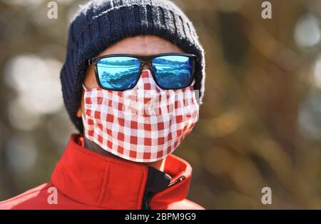 Mann trägt Mütze Hut, blau glänzende Sonnenbrille und weiß rote Baumwolle Hause gemacht Nase Mund Gesicht Virus Maske, Nahaufnahme Detail Porträt, verschwommene Bäume backgr Stockfoto