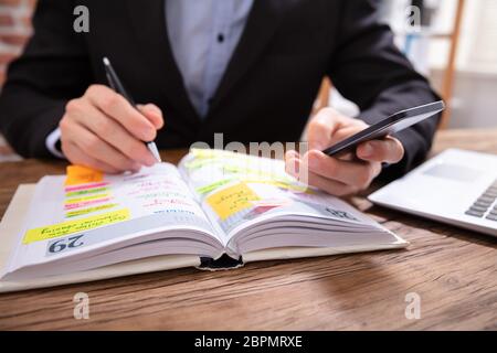 Nahaufnahme eines Kaufmanns Holding Handy Schreiben Zeitplan in das Tagebuch mit Stift Stockfoto