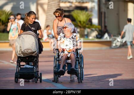 Porto Azzurro, Italien. 26. Juni 2016: Behinderte mit Rollstuhl. Frau mit Kinderwagen. Eine Frau schiebt einen Rollstuhl mit einem Kind hinein. Eine Frau drückt das w Stockfoto