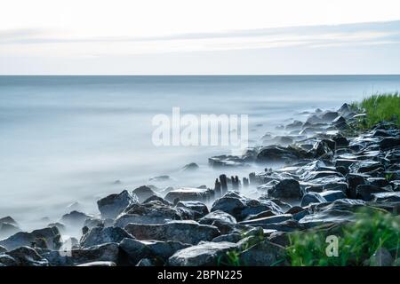 Küste mit Wellen, die auf den Felsen schlagen. Lange Belichtung, verschwommenes Bild von Wasser, Nebel Nebel Nebel zwischen den Felsen am Ufer. Der graue Himmel bedeckt. Stockfoto