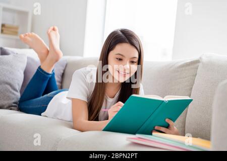 Foto von kleinen hübschen Schüler Schuldame liegen bequem Sofa Lesen Tagebuch Buch positive gute Laune fleißigen Schüler schreiben Notizen Bleistift soziale Distanz Stockfoto