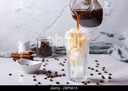 Kalter Kaffee wird in ein hohes Glas mit Eiswürfeln auf hellgrauem Hintergrund gegossen Stockfoto