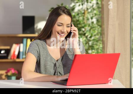 Attraktive Hausfrau mit einem roten Laptop und auf Linie mit einem Mobiltelefon in einer Tabelle in einem Haus-Interieur in den Kundendienst anrufen Stockfoto