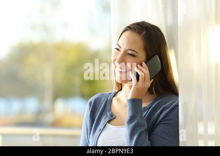 Glückliche Frau Gespräch am Handy stand neben einem Fenster zu Hause Stockfoto