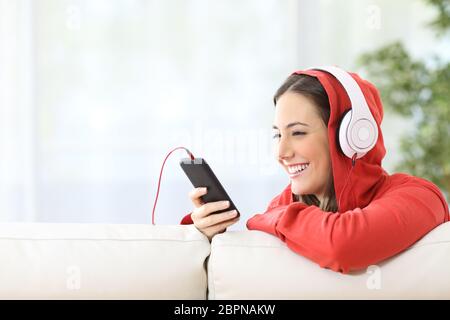 Porträt eines glücklichen Teenagers Musik online von Smartphone hören sitzen in einer Couch zu Hause mit weißen Vorhängen im Hintergrund Stockfoto