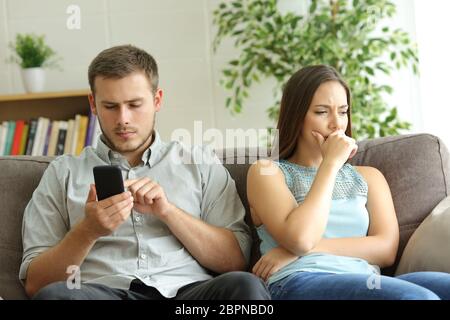Ehemann zu smart phone Sie Inhalt neben seinem besorgt, Frau, sitzen auf einem Sofa zu Hause süchtig Stockfoto