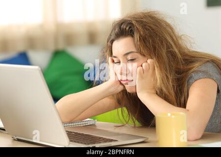 Verschlafene Schüler mit zerzausten Haaren versucht zu lesen, auf Linie Inhalt in einem Laptop auf dem Schreibtisch in ihrem Zimmer in einem Haus-Interieur Stockfoto